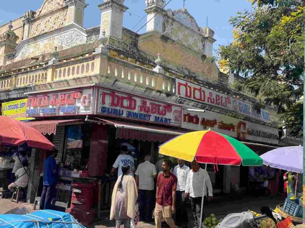 Guru Sweet Mart in Devaraja Mohalla, Mysuru, showing the busy traditional sweet shop with customers and street market surroundings