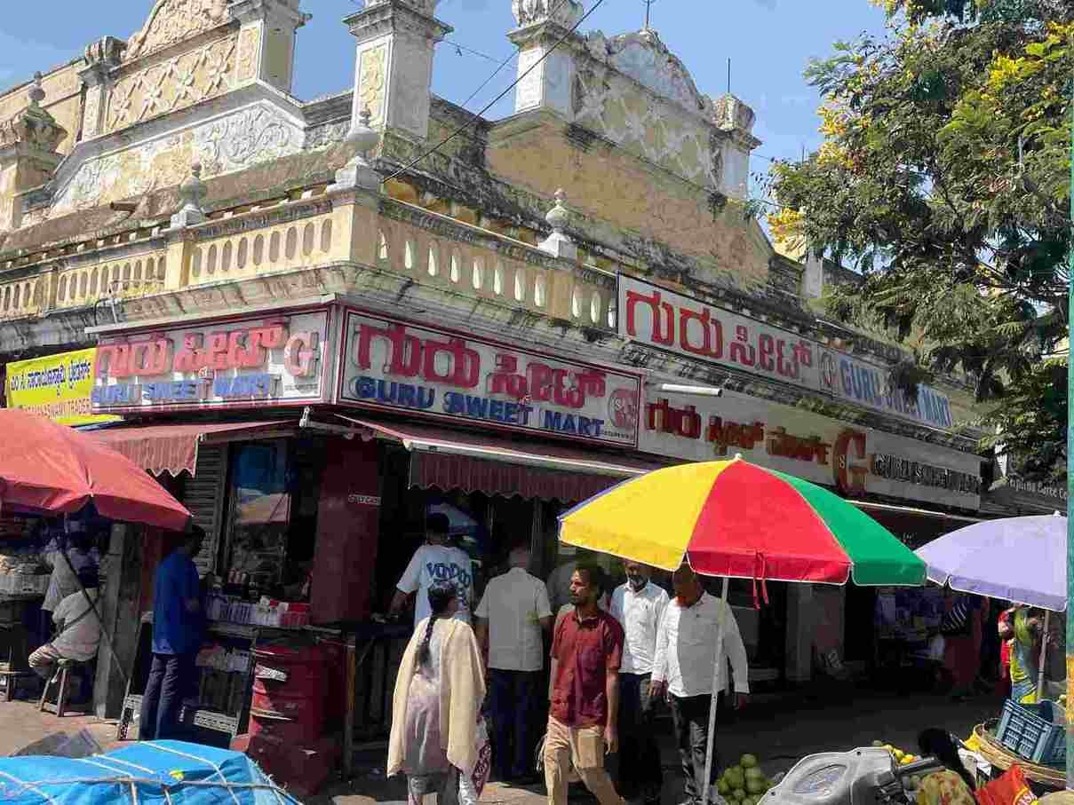 Guru Sweet Mart in Devaraja Mohalla, Mysuru, showing the busy traditional sweet shop with customers and street market surroundings
