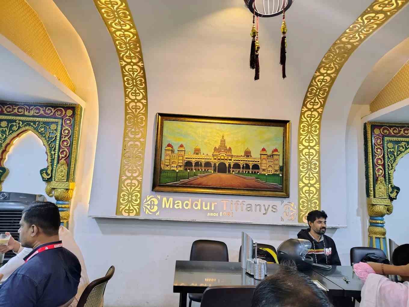 Elegant interior seating area of Maddur Tiffanys Mysore with Mysore Palace artwork and traditional Karnataka-inspired décor.