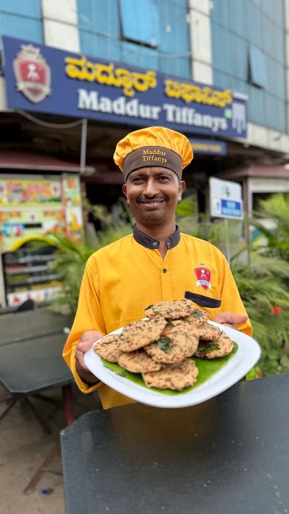 Staff member at Maddur Tiffanys holding a plate of freshly prepared Maddur Vadas outside the restaurant.